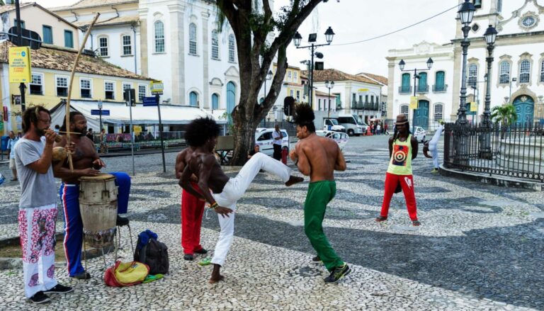 Roda de capoeira em uma praça histórica do Pelourinho, mostrando homens tocando berimbau e atabaque à esquerda, enquanto dois capoeiristas jogam ao centro sobre o calçamento de pedras.